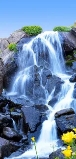 Stunning waterfall amidst rocky terrain with vibrant yellow flowers and blue sky.