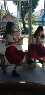 Two school girls seated outside using smartphones in the sunlight.