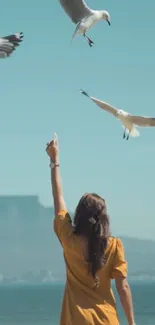 A person in a golden dress points at flying seagulls by the ocean.