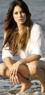 Woman crouching by the beach, wearing white blouse and shorts.