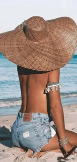 Woman in sun hat sitting on sandy beach by ocean.