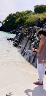 Person standing on a serene beach with rocky shoreline and green trees.