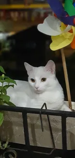 White cat resting beside a colorful windmill and flower pot in a serene setting.