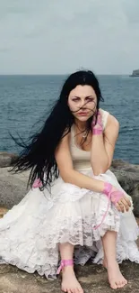 Woman in white dress sitting on rocks by the ocean with a serene expression.