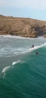 Two surfers ride waves near a rocky coast under a clear sky.