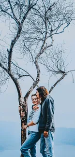 Couple embracing under a bare tree with a blue sky backdrop.