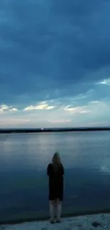 Person standing by a calm lakeside at dusk with a moody blue sky.