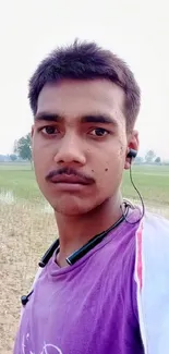 Young man in purple shirt with earphones standing in serene field.