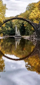 Stone arch bridge reflecting on a calm forest lake with autumn foliage.