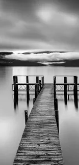 Monochrome image of a wooden pier extending into a serene lake under a cloudy sky.