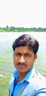 Man taking a selfie by a green lakeside with clear blue sky.