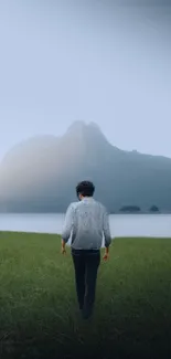 Solitary figure by a misty lake with mountains and green field.