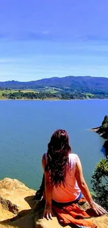 Person sitting by a tranquil lake with mountains and blue sky.