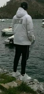 Person in white jacket by a serene lakeside with boats.