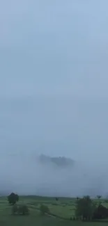 Misty landscape with green fields and fog under a blue sky.