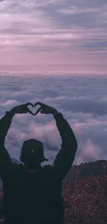 Silhouette with heart hands against pink sunset mountain and clouds.