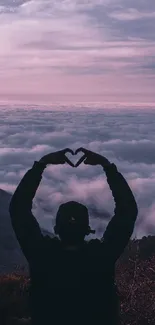 Silhouette of person forming heart hands against pink sunset and mountain clouds.