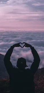 Silhouette of person forming heart with hands against sunset over mountains and clouds.