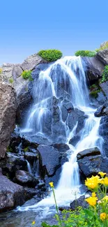 Serene mountain waterfall with vibrant flowers and a clear blue sky.