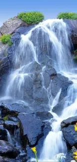 A serene mountain waterfall with vibrant yellow flowers and lush greenery.