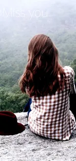 Person in a checkered shirt sits in nature, gazing at misty forest.