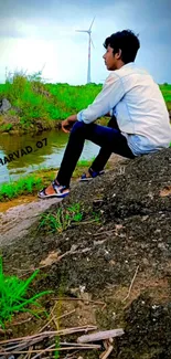 Man sitting on rock by river with green fields and wind turbine.