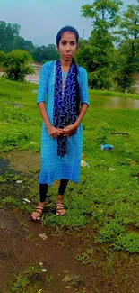 Woman standing outdoors in blue dress with green landscape background.