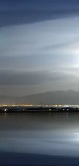 Serene night sky reflecting on a calm lake with lights in the horizon.