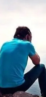 Person in blue shirt sits contemplatively by the ocean's edge.