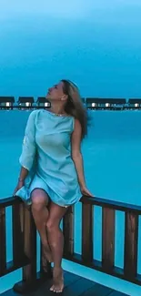 Woman sitting by the ocean in a turquoise dress, gazing at tranquil waters.