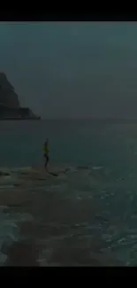 Person on rock by the serene teal ocean with mountain backdrop.