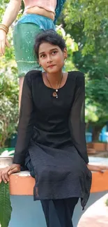 Young woman in black dress sitting outdoors with green leafy background.