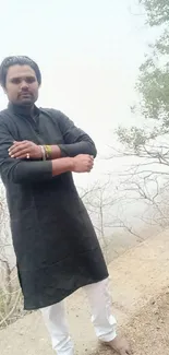 Man in black attire standing outdoors with a natural backdrop.