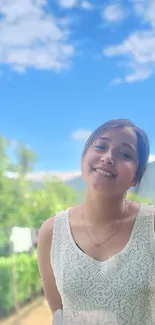 Woman smiling outdoors with blue sky and lush greenery backdrop.