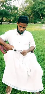 A person in white clothing sitting outdoors on grass, reading.