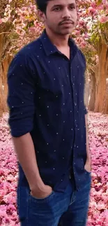 A man standing on a path covered in pink blossoms under tall trees.