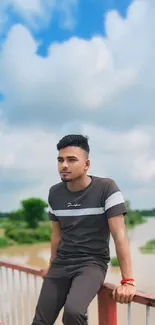 Young man sitting on a bridge against a blue sky and tranquil river landscape.