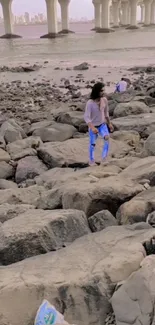 Woman walking along a rocky beach with a city view.