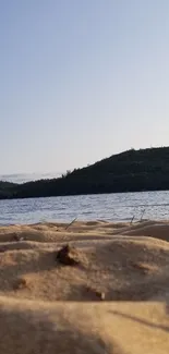 Sandy beach with lake and forested hills under clear sky.