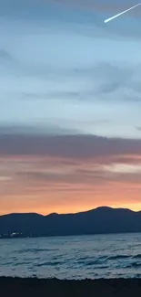 Serene beach at sunset with mountain silhouettes and a meteor in the sky.