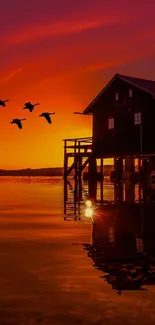 Sunset over lake with house, birds flying in vibrant orange sky.