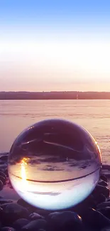 Glass sphere on pebbles reflecting a serene sunset over the ocean.