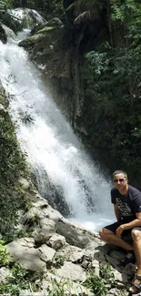 Man sitting near a lush waterfall with greenery surrounding.