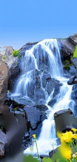 Serene waterfall running over rocks with bright blue sky and yellow flowers.