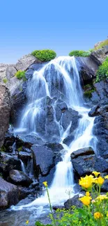 A serene waterfall surrounded by rocks and vibrant flowers under a clear blue sky.