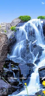 Serene waterfall with rocks and yellow flowers under a blue sky.