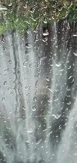 Waterfall seen through raindrop-covered glass, lush green backdrop.