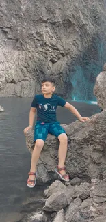 Boy sitting on rocks by a serene waterfall amidst nature.