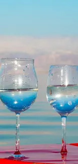 Two wine glasses reflecting calm waters and a blue sky in serene setting.