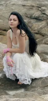 Woman in white dress sits on rocky shore, looking serene and tranquil.